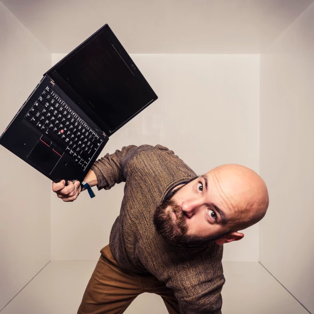 Christoph Döberl, Mann mit rasiertem Kopf und Bart, hält scherzhaft einen aufgeklappten Laptop über den Kopf
